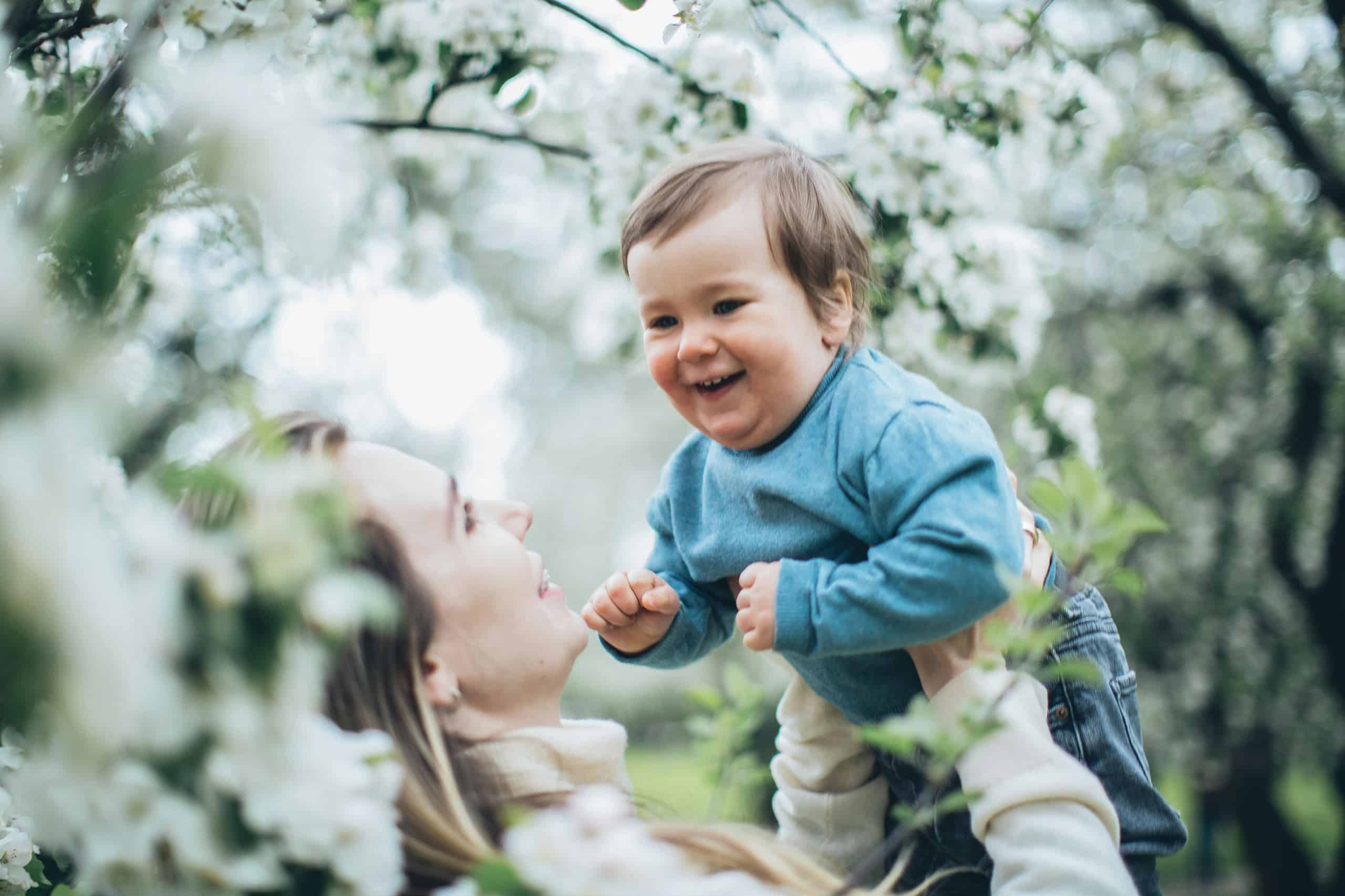 Nanny and baby (in a blue sweater) close together outdoors with soft greenery in the background