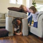 Nanny playing hide and seek with two giggling girls under couch cushions, joy in every visit