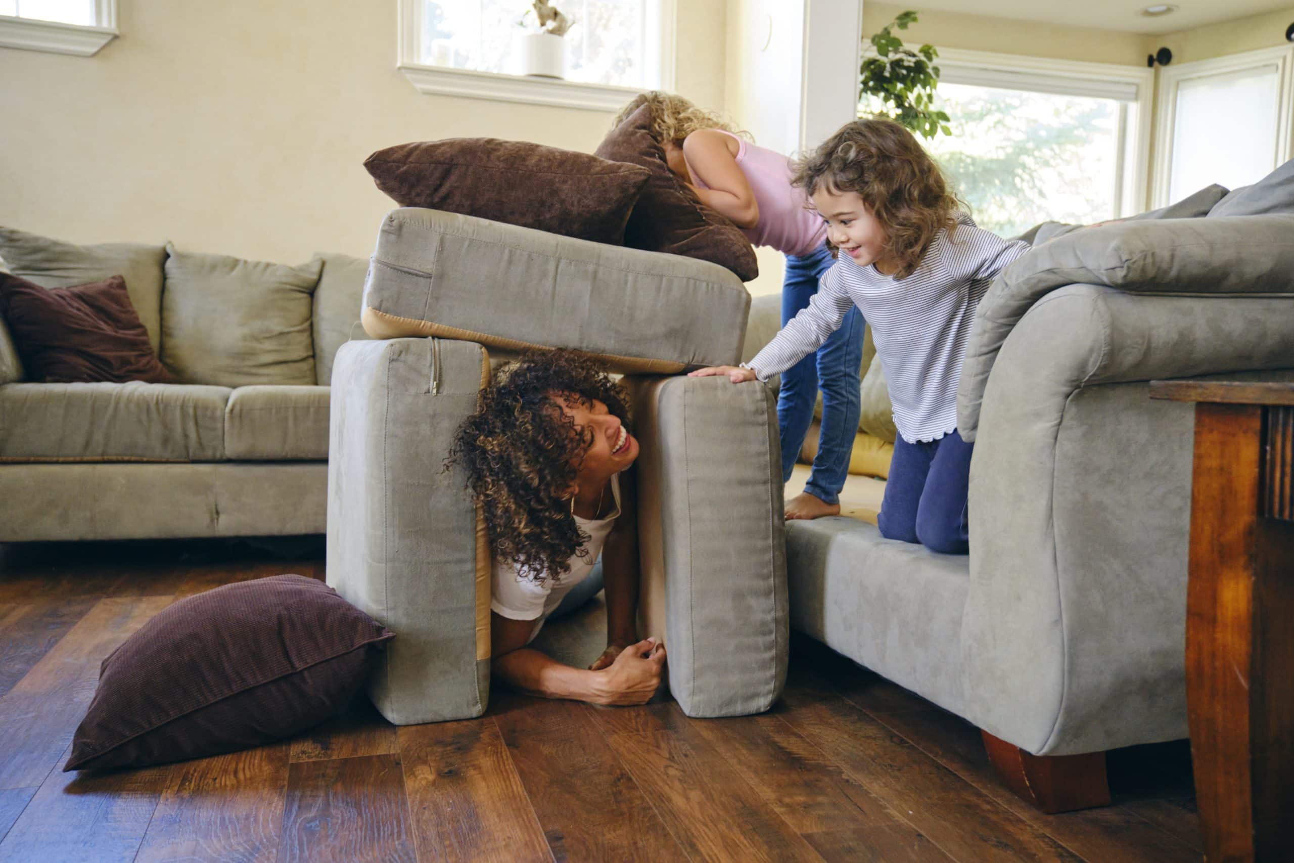 Nanny playing hide and seek with two giggling girls under couch cushions, joy in every visit