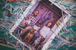A physical photo print of a smiling couple with two dogs and a cat, lying on a rustic teal wooden surface amidst a large collection of colorful capped medical syringes. A physical photo print of a smiling couple with two dogs and a cat, lying on a rustic teal wooden surface amidst a large collection of colorful capped medical syringes.